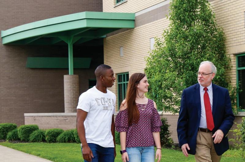 三个人 are walking and conversing outside a building with green trim. A man in a suit and tie is accompanied by two young adults, one in a patterned blouse and jeans, and the other in a white T-shirt.